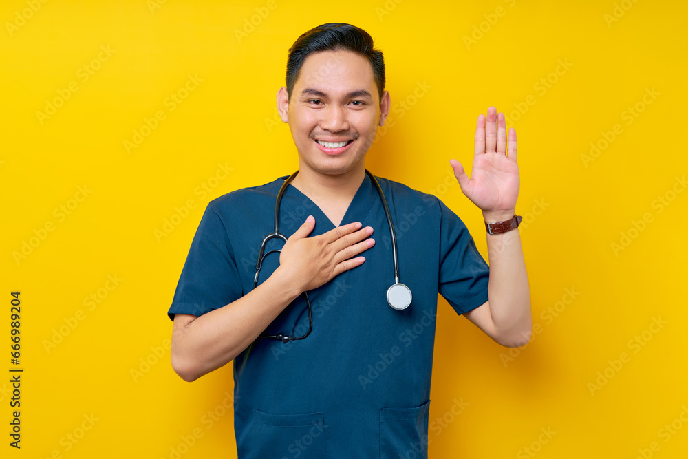Smiling professional young Asian male doctor or nurse wearing a blue uniform and stethoscope waving hand to say hi, hello, greeting patient, hand on chest isolated on yellow background Smiling professional young Asian male doctor or nurse wearing a blue uniform and stethoscope waving hand to say hi, hello, greeting patient, hand on chest isolated on yellow background
