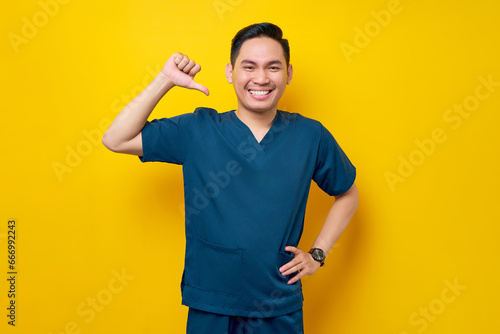 Smiling professional young Asian male doctor or nurse wearing a blue uniform standing confident while pointing a finger at himself isolated on yellow background. Healthcare medicine concept