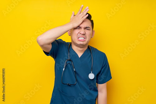 Mistake professional young Asian male doctor or nurse wearing a blue uniform looking at camera and hand on his head isolated on yellow background. Healthcare medicine concept