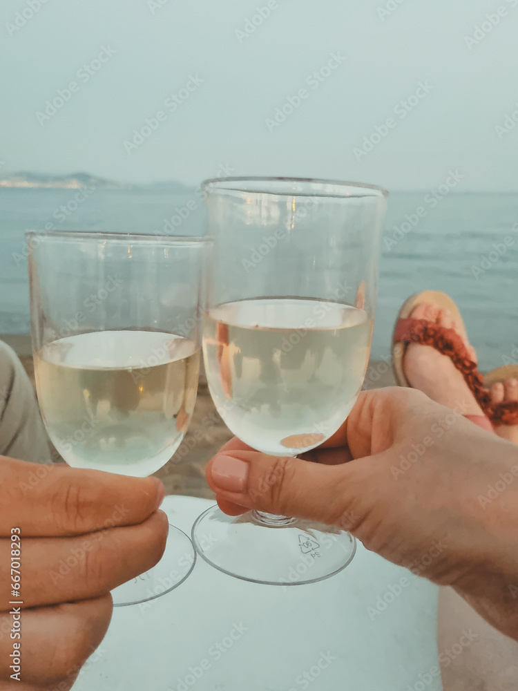 Obraz premium female and male hands hold glasses of white wine on a sandy beach against the backdrop of the sea. summer vacation and beach holiday concept. soft focus. vertical photo