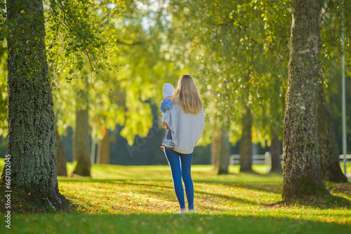 Fotografie Young adult mother carrying baby boy in arms at tree alley at city park