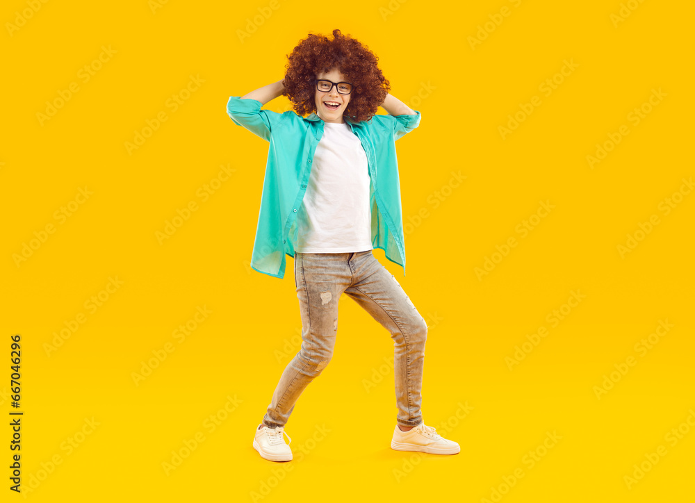 Happy funny preteen boy in curly wig dancing and having fun. Full size photo of joyful boy wearing glasses, turquoise shirt, jeans and sneakers standing against yellow studio background