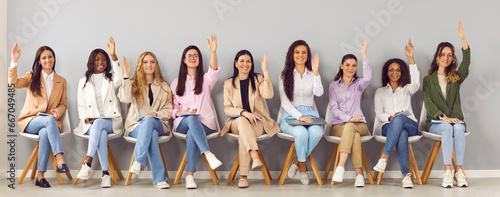 Portrait of group of happy diverse business women company employees looking at camera and raising hands to ask or answer a question sitting in a row on meeting on a gray wall background. Banner. photo