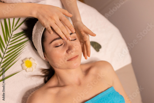 Spa And Relax. Young Lady Receiving Head Massage Enjoying Beauty Ritual Lying With Eyes Closed On Bed Indoor, Top View. Masseur Massaging Lady's Forehead And Face For Relaxation