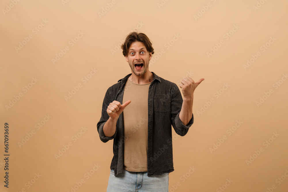 Excited man pointing fingers away at copy space while standing isolated over beige wall Excited man pointing fingers away at copy space while standing isolated over beige wall