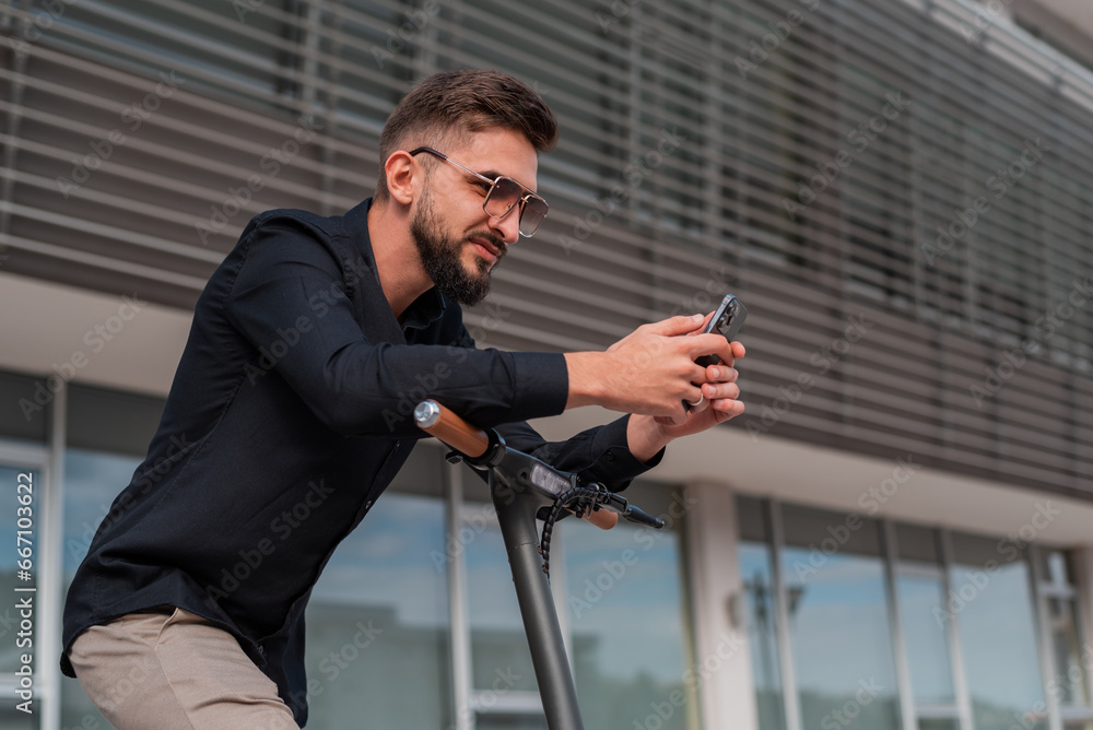 A young happy man with electric scooter standing in front of modern business building on city street and using a phone. A young happy man with electric scooter standing in front of modern business building on city street and using a phone.