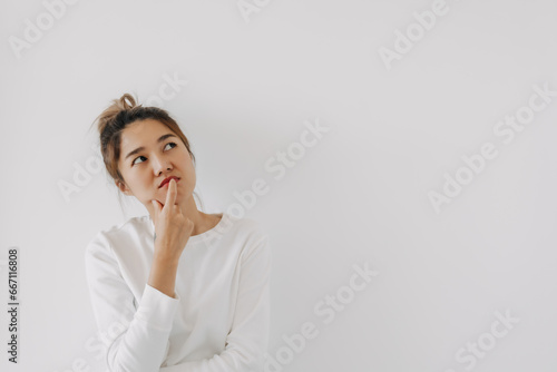 Asian Thai woman wear sweater bun hair, touching chin and mouth with doubt and curious face, have question thinking and looking at empty space, isolated on white background wall at winter. photo