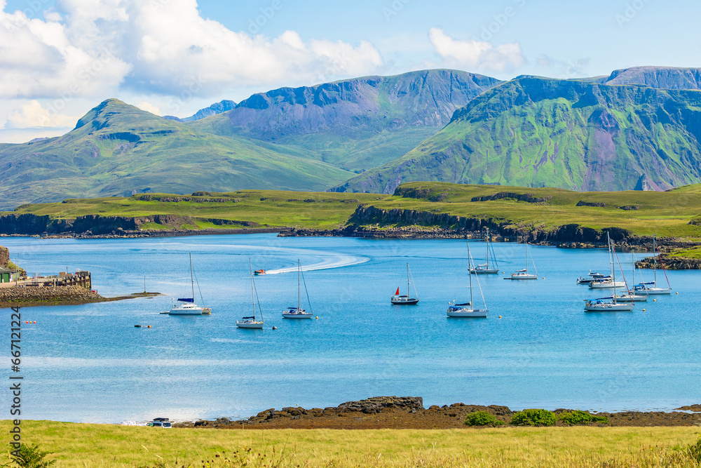 The beautiful Isle of Canna, Inner Hebrides in Summer with yachts and a catamaran in the bay and a RIB speeding towards the harbour with the Isle of Rum in the background.  Horizontal.  Copy space - obrazy, fototapety, plakaty 