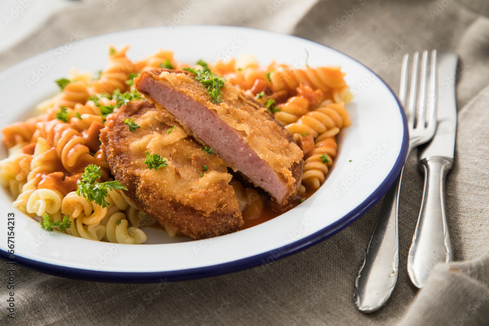 Jägerschnitzel nach DDR Rezept mit Jagdwurst paniert, Spirelli Fusilli Nudeln, Tomatensoße, Petersilie auf Emaille Teller, Besteck und grauer Leinen Serviette Jägerschnitzel nach DDR Rezept mit Jagdwurst paniert, Spirelli Fusilli Nudeln, Tomatensoße, Petersilie auf Emaille Teller, Besteck und grauer Leinen Serviette