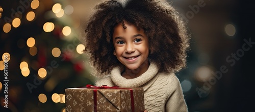 A cheerful African American girl sits on Santa s lap near a festive Christmas tree opening a gift and expressing joy and surprise photo