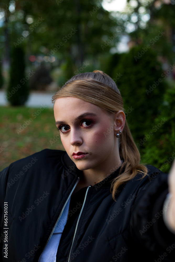 European blonde girl with a ponytail with bright makeup in a black bomber jacket posing taking a selfie with her phone in the park. Vertical photo of a portrait of a made-up girl looking at the camera