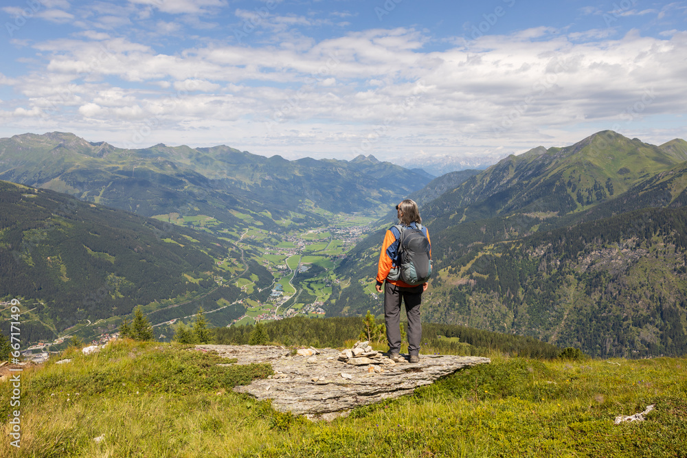 Gray-haired long-haired male hiker with backpack standing at the edge of cliff and looking at a mountain valley, Austria