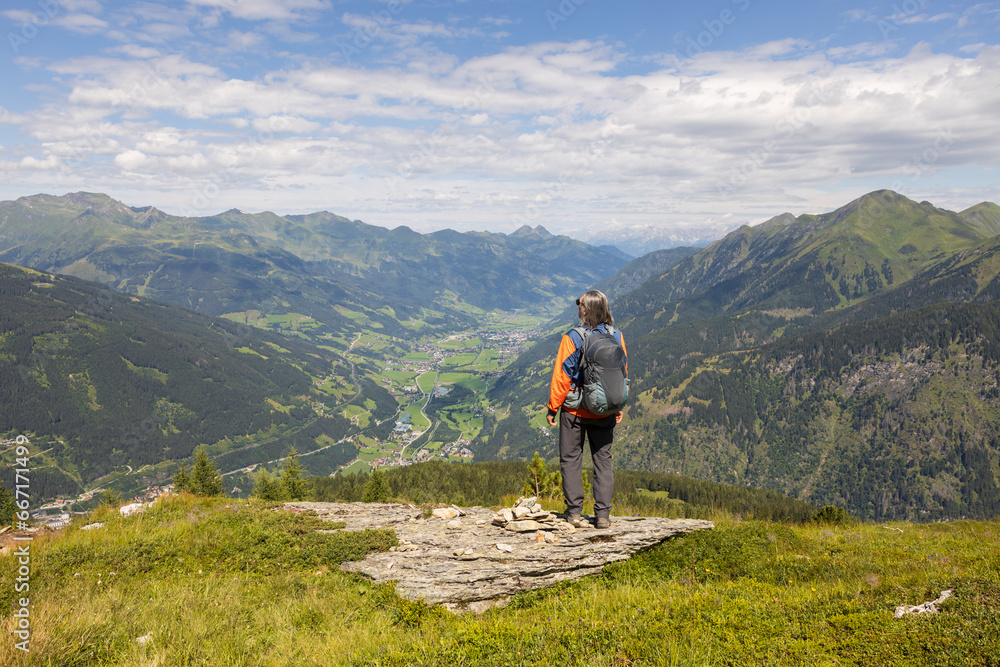 Gray-haired long-haired male hiker with backpack standing at the edge of cliff and looking at a mountain valley, Austria Gray-haired long-haired male hiker with backpack standing at the edge of cliff and looking at a mountain valley, Austria