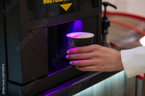 Close-up view of female hand taking out black disposable paper coffee cup with latte coffee drink from table top vending coffee machine in micro market. Soft focus. Self service theme.