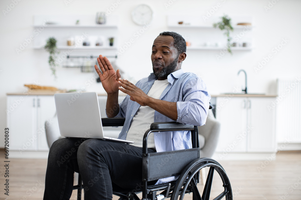Young african american person in casual wear with portable computer while sitting in wheelchair in studio apartment. Positive adult man having video call, while sitting at kitchen. - obrazy, fototapety, plakaty 