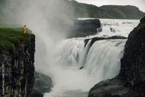 Picture of a waterfall in Iceland with a man in a yellow raincoat standing on a cliff and enjoying the wild nature.