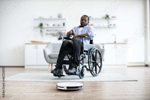 Focus on smart robotic vacuum operated by happy african man with mobility impairment while performing automatic cleaning on carpet indoors. Cheerful owner programming home gadget via remote control.