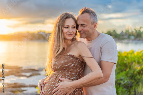 A happy, mature couple over 40, enjoying a leisurely walk on the waterfront On the Sunset, their joy evident as they embrace the journey of pregnancy later in life