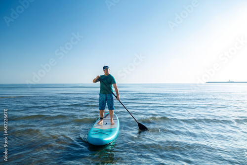 Fotografie A man in shorts and a T-shirt stands on a SUP board with a paddle near the seashore in the morning at dawn