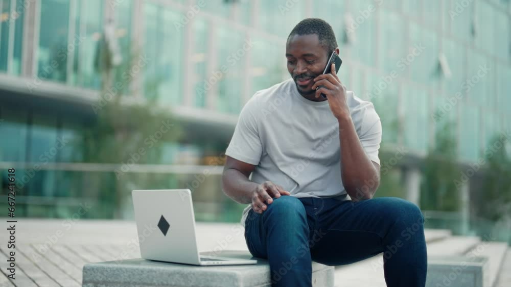 Man entrepreneur calling on smartphone looking at laptop screen sitting outdoors near office building in city downtown outdoors. Positive professional guy freelancer working, having conversation. Man entrepreneur calling on smartphone looking at laptop screen sitting outdoors near office building in city downtown outdoors. Positive professional guy freelancer working, having conversation.