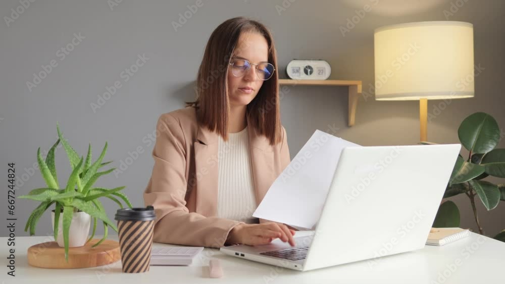 Young successful concentrated employee business woman in official style jacket doing paper documents work at workplace sitting at desk with laptop pc computer at light modern office indoors. Young successful concentrated employee business woman in official style jacket doing paper documents work at workplace sitting at desk with laptop pc computer at light modern office indoors.