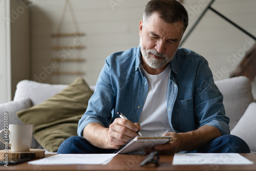 Senior man taking notes in notebook while sitting on the couch, gray-haired elderly man writing thoughts in notebook