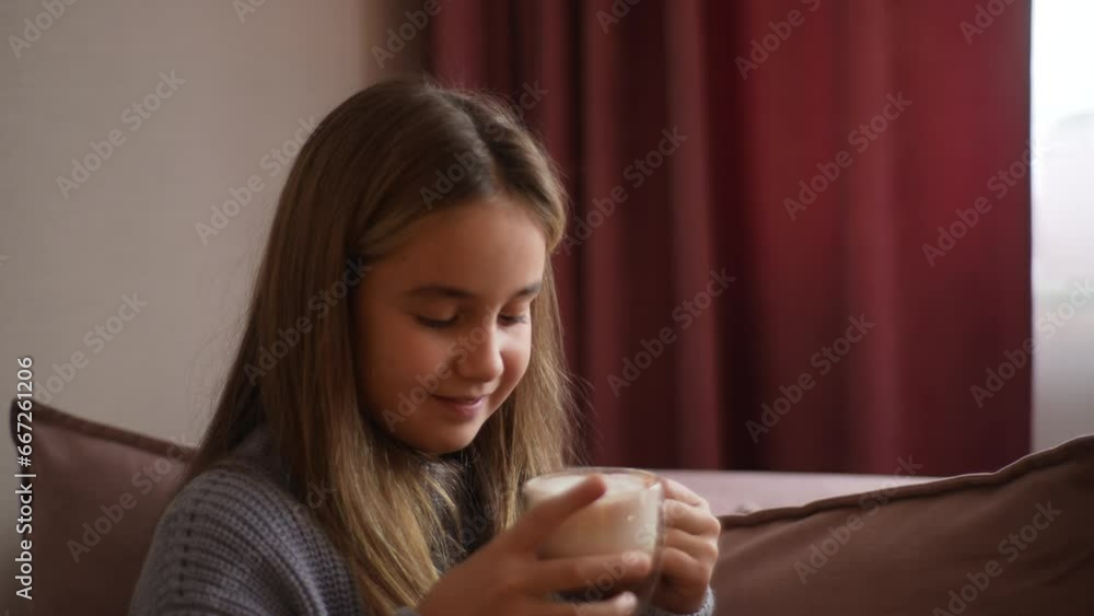 Portrait of smiling cute little girl drinking hot cocoa from mug sitting on sofa at home. Closeup of cheerful adorable kid drinking tasty chocolate cocktail with happy expression, slow motion. Portrait of smiling cute little girl drinking hot cocoa from mug sitting on sofa at home. Closeup of cheerful adorable kid drinking tasty chocolate cocktail with happy expression, slow motion.