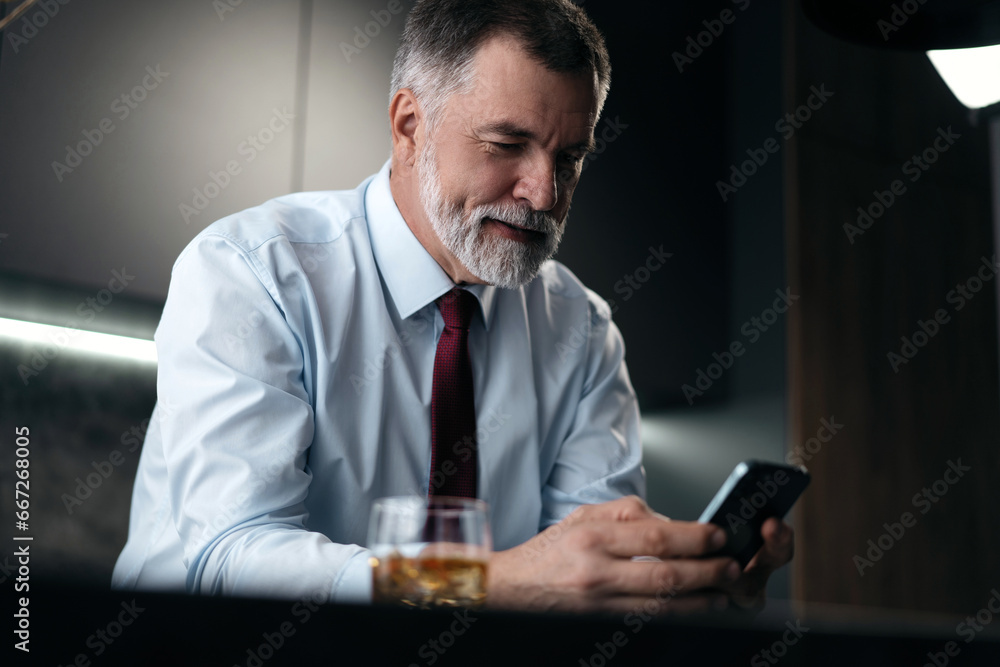 Mature businessman with glass of whiskey using smartphone while sitting in the kitchen. Mature businessman with glass of whiskey using smartphone while sitting in the kitchen.