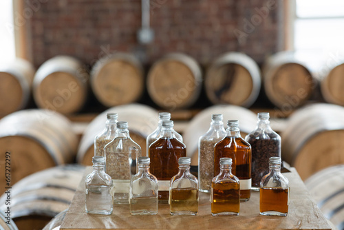 collection of clear glass bottles in different sizes filled with whiskey and bourbon sitting as display surrounded by whiskey barrels in distillery