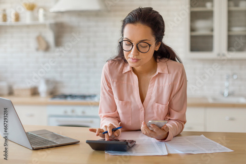 Focused woman with laptop, cellphone and calculator in kitchen