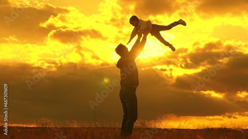 Fototapeta Naklejka Na Ścianę i Meble -  Happy dad plays with his baby daughter, throws child into sky with his hands, happy child laughs. Silhouette, father, child playing together in park against backdrop of sun clouds. Family, dream fly