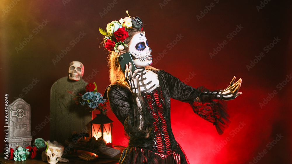 Young scary woman talking on smartphone call, wearing halloween make up and flowers crown in studio. La cavalera catrina using smartphone to chat on dios de los muertos mexican holiday.