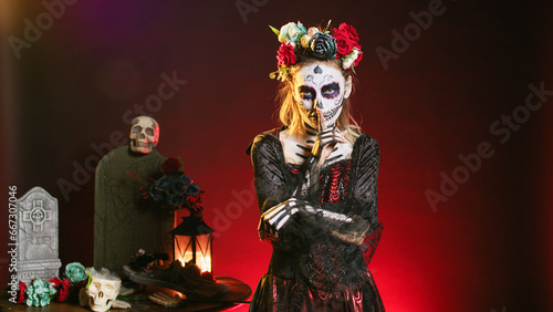 Creepy female model doing hush gesture to keep secret, wearing santa muerte costume and showing slience mute and secrecy sign with finger over lips. Holy dios de los muertos holiday.