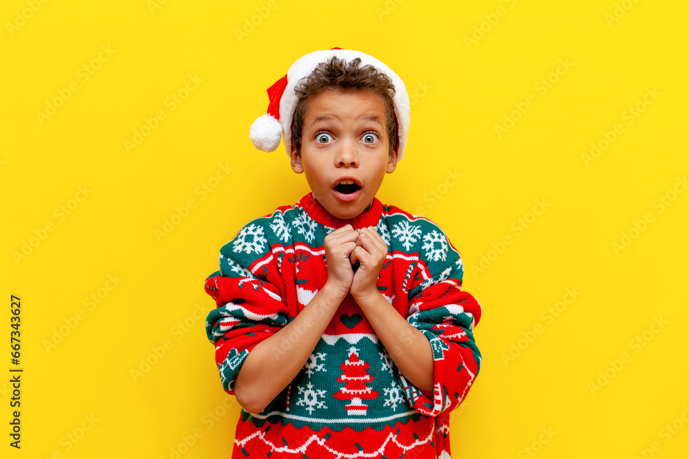 shocked african american boy in christmas sweater and santa hat looking surprised on yellow isolated background