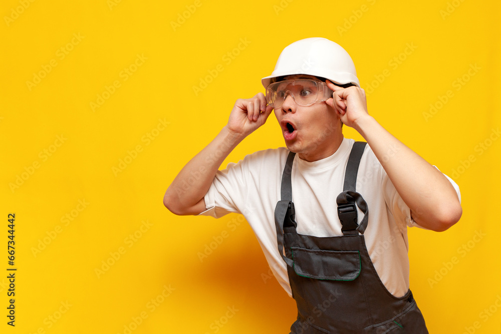 shocked asian male construction worker in uniform and safety glasses looking in amazement over yellow background shocked asian male construction worker in uniform and safety glasses looking in amazement over yellow background