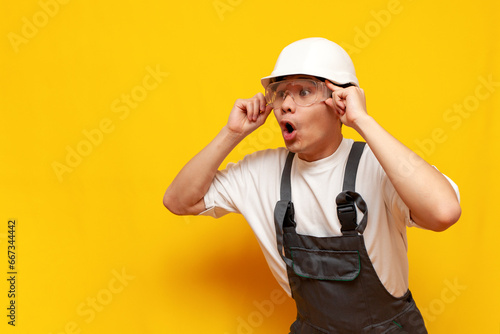 shocked asian male construction worker in uniform and safety glasses looking in amazement over yellow background