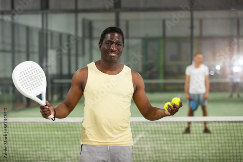 Portrait of cheerful confident african american paddle tennis player standing on indoor court during friendly match © JackF