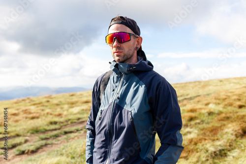 young man in jacket and sunglasses stands on the mountain and looks at the sun, tourist guy in mountain clothes
