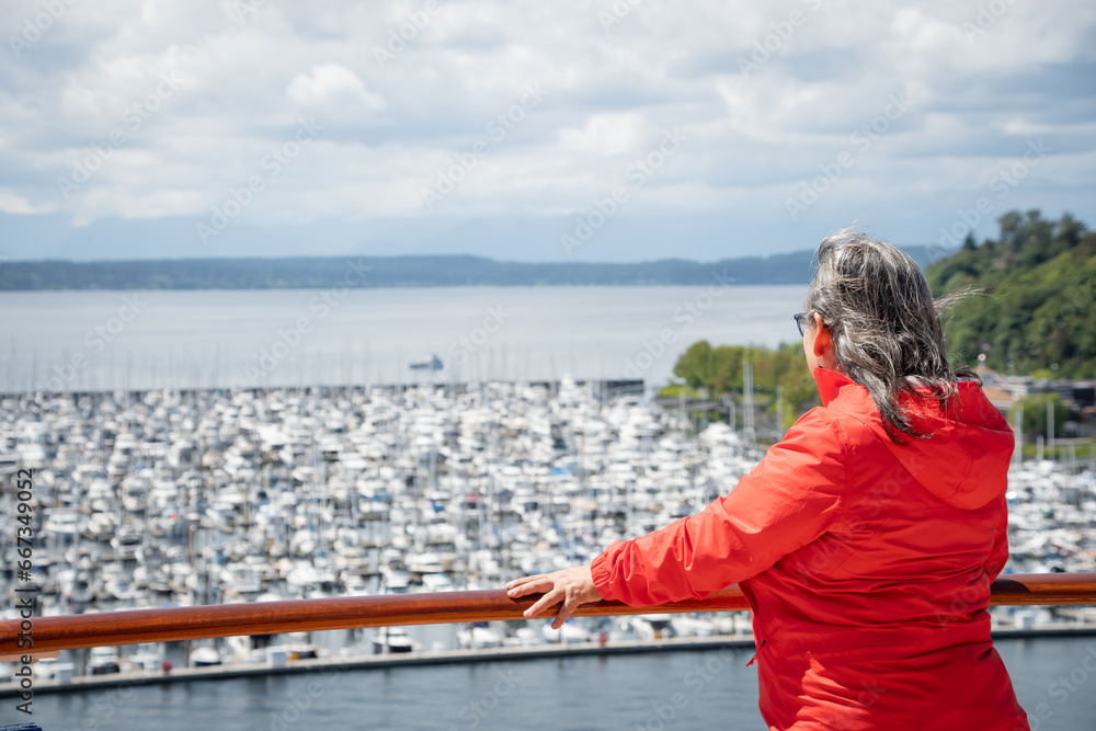 Woman with her back to the camera, wearing an orange windbreaker, gazes at the horizon from a marina.