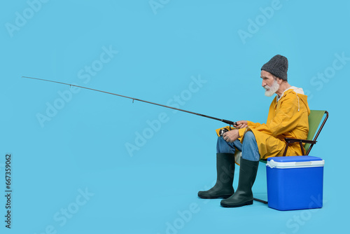 Fisherman with rod and cool box on chair against light blue background
