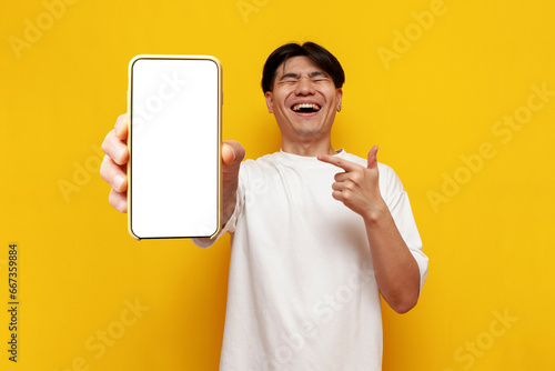 young asian guy in glasses shows blank screen of smartphone and laughs on yellow isolated background