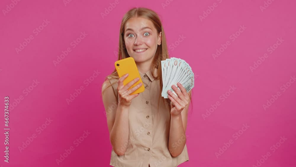 Young woman looking smartphone display sincerely rejoicing win, receiving money dollar cash banknotes, celebrating success lottery luck. Girl isolated alone on pink studio background. Lifestyles Young woman looking smartphone display sincerely rejoicing win, receiving money dollar cash banknotes, celebrating success lottery luck. Girl isolated alone on pink studio background. Lifestyles