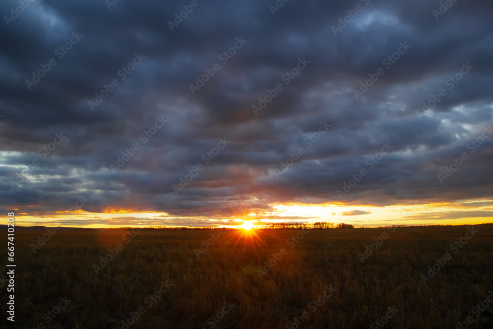 Sunset with bright orange rays and cloudy sky over the field.