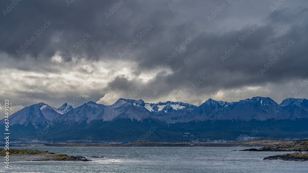 Beautiful snow-capped mountain range of the Andes against a cloudy sky. The city houses of Ushuaia are visible at the foot. In the foreground are islands in the Beagle Channel. Argentina. Beautiful snow-capped mountain range of the Andes against a cloudy sky. The city houses of Ushuaia are visible at the foot. In the foreground are islands in the Beagle Channel. Argentina.