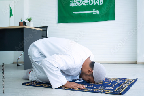Side view of Arab Muslim man doing Salat on prayer mat at office, Saudi Arabia Flag hanging on wall in background