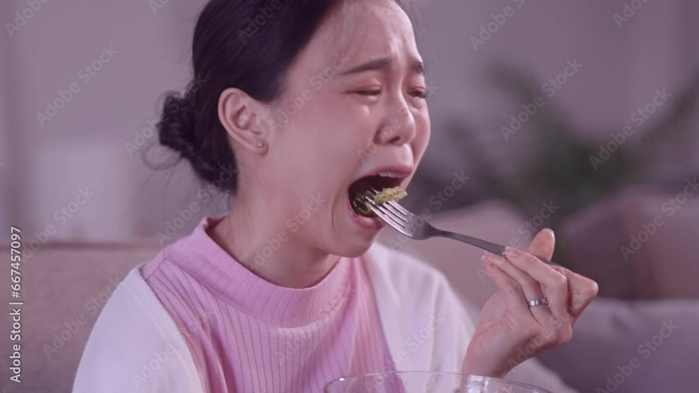 Asian teenage girl is reluctantly eating a vegetable salad from a glass bowl, even though she doesn't like vegetables. She's seated on her living room sofa at her own home to be healthy Asian teenage girl is reluctantly eating a vegetable salad from a glass bowl, even though she doesn't like vegetables. She's seated on her living room sofa at her own home to be healthy