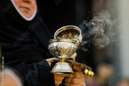 Shallow depth of field (selective focus) details with the hand of a orthodox Christian nun holding a smoking metallic censer incense burner.