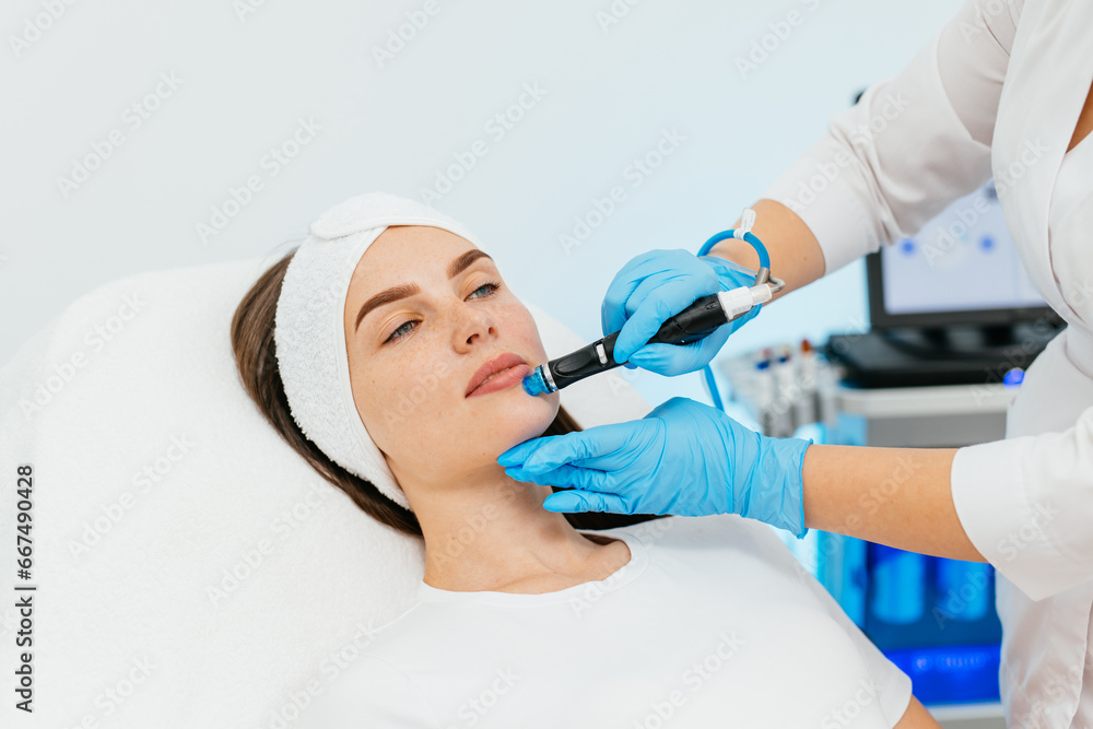 Fototapeta premium Young woman receiving facial treatment in a professional beauty salon. A close-up cosmetologist manipulates a hydropeeling machine to clean and rejuvenate a patient's skin in a modern clinic.