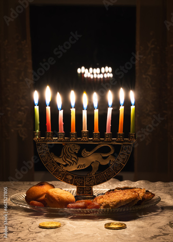 Vertical view of Menorah with candles lit for Jewish holiday Hanukkah with traditional sweets; candles reflect in dark window in background photo