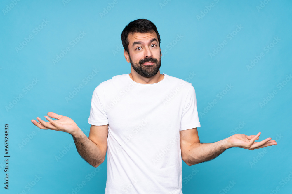 Young man with beard  over isolated blue background having doubts while raising hands